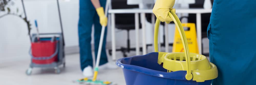 Close-up of a cleaning professional holding a mop bucket in an office space with another cleaner mopping the floor in the background.